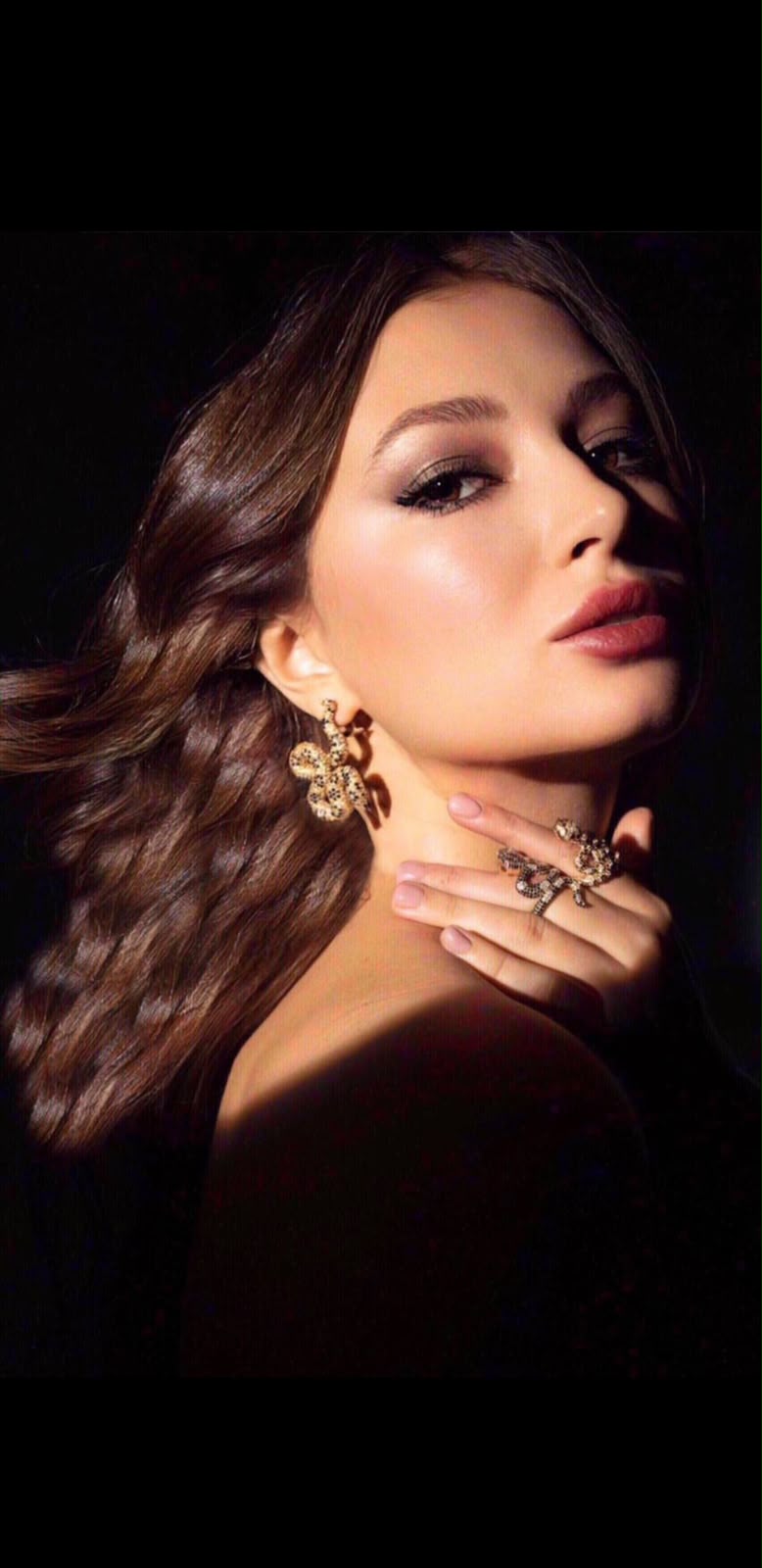 Close beauty portrait of Lilia Mai wearing statement gold earrings and a jewelled ring, against a dark backdrop.