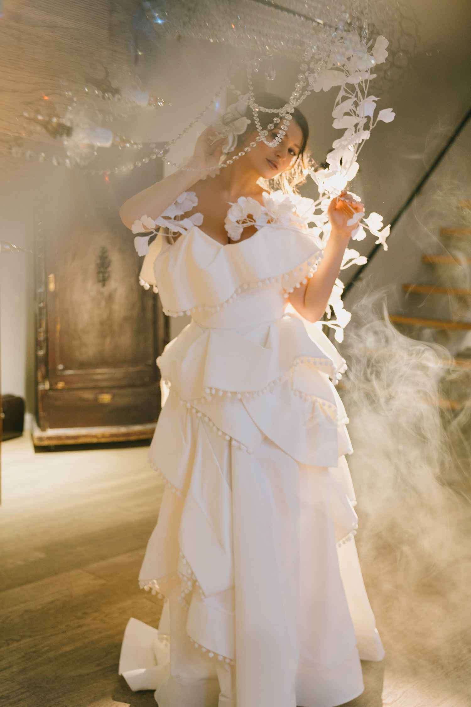 Full-length view of Lilia Mai in a tiered white couture gown, lit through smoke beneath a crystal chandelier.